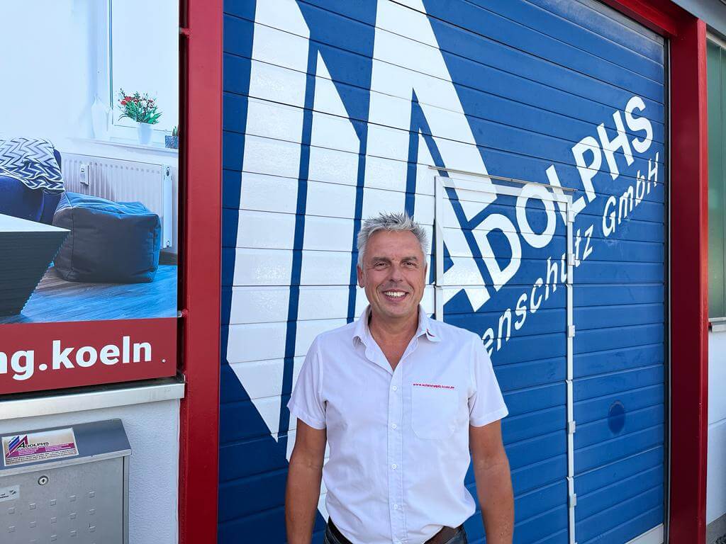 Marco Bongartz steht im weißen Hemd vor der Lagerhalle von Adolphs Bautenschutz und lacht